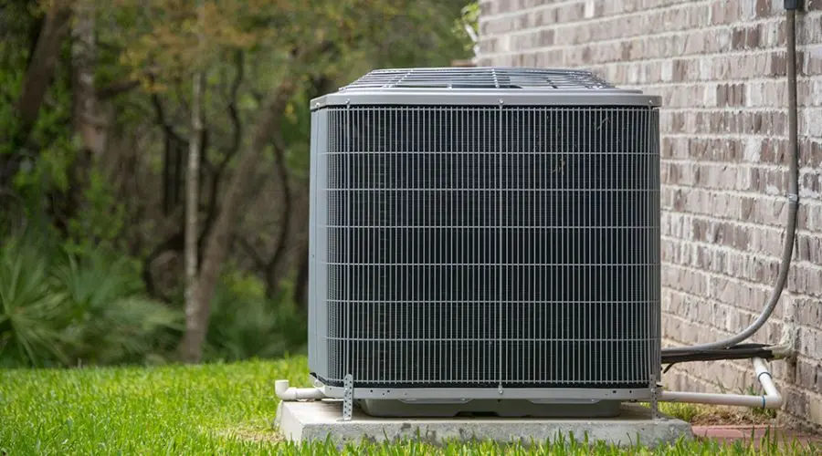 Air conditioning unit on a concrete base against an exterior wall, surrounded by a well-maintained grass lawn.
