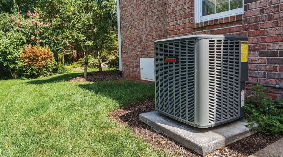 Air conditioning unit near brick building on concrete base surrounded by shrubs and grass, reflecting City Plumbing services.