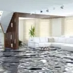 Flooded living room with water covering the floor, featuring a white sectional sofa, wooden accents, and natural light from large windows, highlighting water damage risks discussed by City Plumbing.
