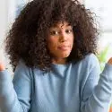 Woman with curly hair in light blue sweater, unsure, in a bright indoor setting related to plumbing services.
