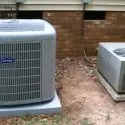 Two air conditioning units on concrete pads, vital for HVAC maintenance by City Plumbing. Gray casings by a brick wall.