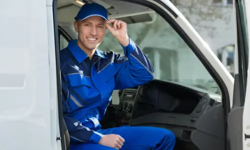HVAC technician in blue uniform and cap smiling from inside service van, representing emergency heating repair services.