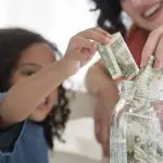 Child and adult placing cash into a clear jar, representing savings and financial education.