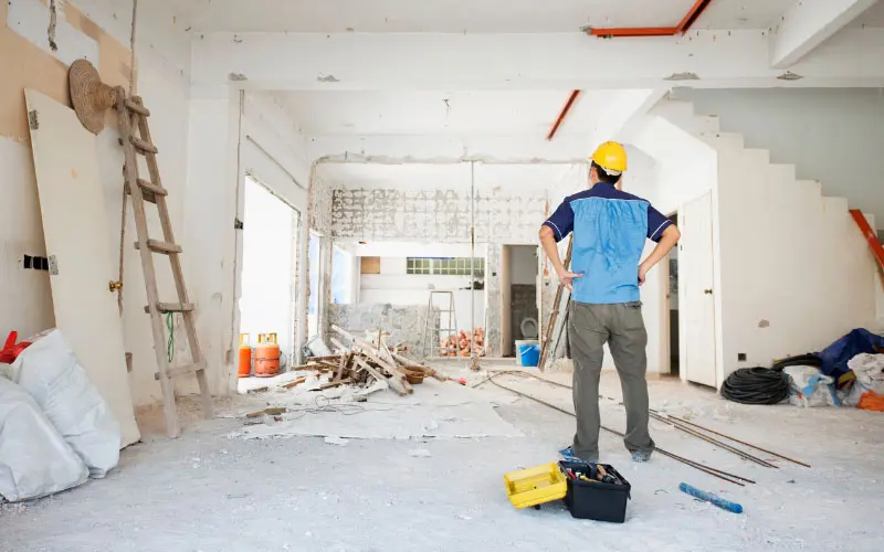 Construction worker assessing a home renovation project in an unfinished interior space, surrounded by tools and debris, emphasizing preparation for holiday upgrades.