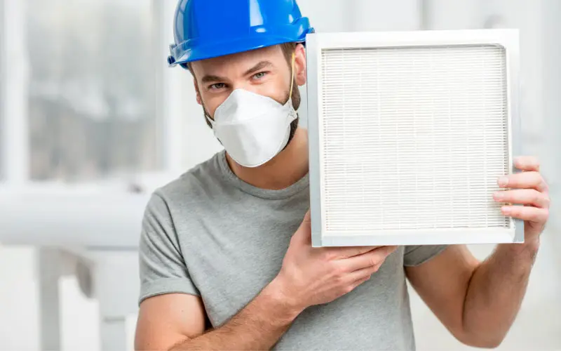 Man in a blue hard hat and mask holding an air filter, emphasizing HVAC maintenance and air quality importance.