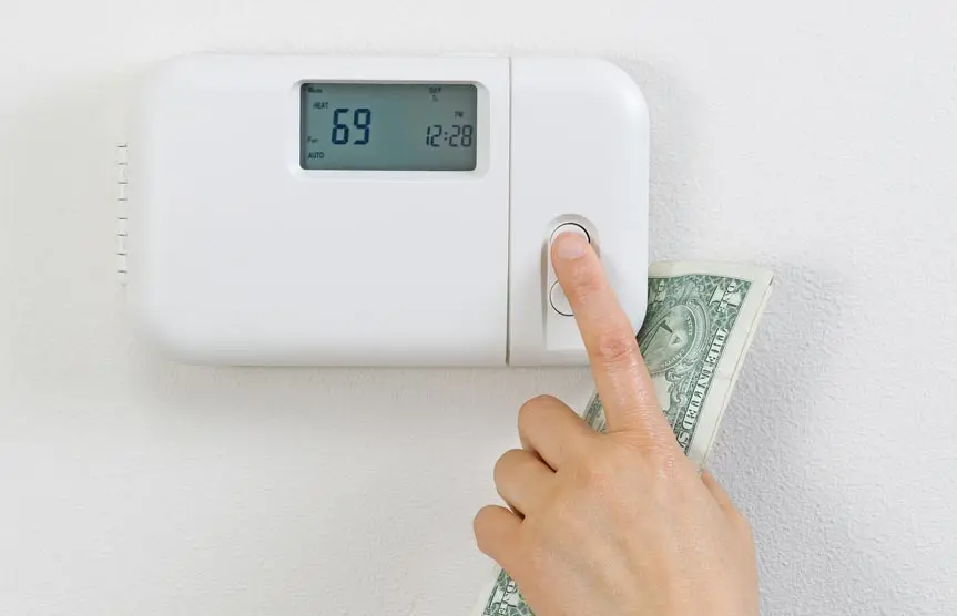 Hand adjusting a thermostat displaying 69°F while holding a dollar bill, symbolizing HVAC efficiency and cost management for home comfort.