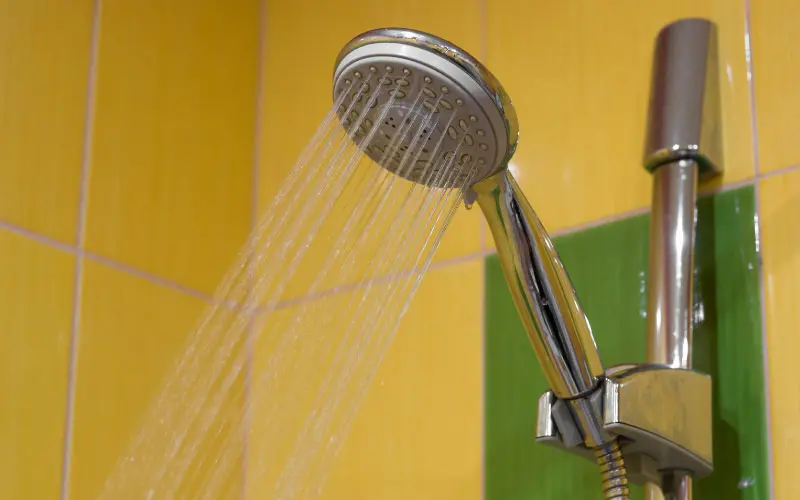 Showerhead spraying water against a vibrant yellow and green tiled wall, illustrating water pressure issues in plumbing.