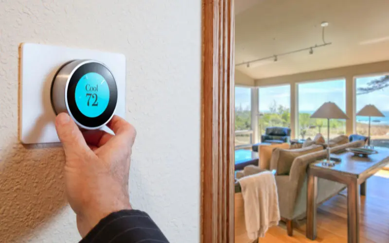Hand adjusting a smart thermostat displaying "Cool 72" in a modern living room setting, illustrating the benefits of smart home technology for HVAC efficiency.