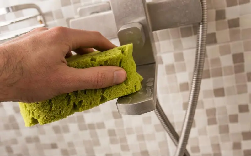 Hand using a green sponge to clean a faucet, demonstrating maintenance to prevent hard water stains in a bathroom setting.