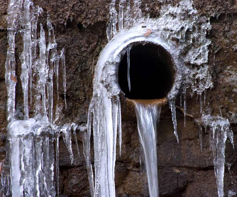 Frozen pipe with icicles and water dripping from a black drainage hole, illustrating winter plumbing issues and the importance of pipe protection.