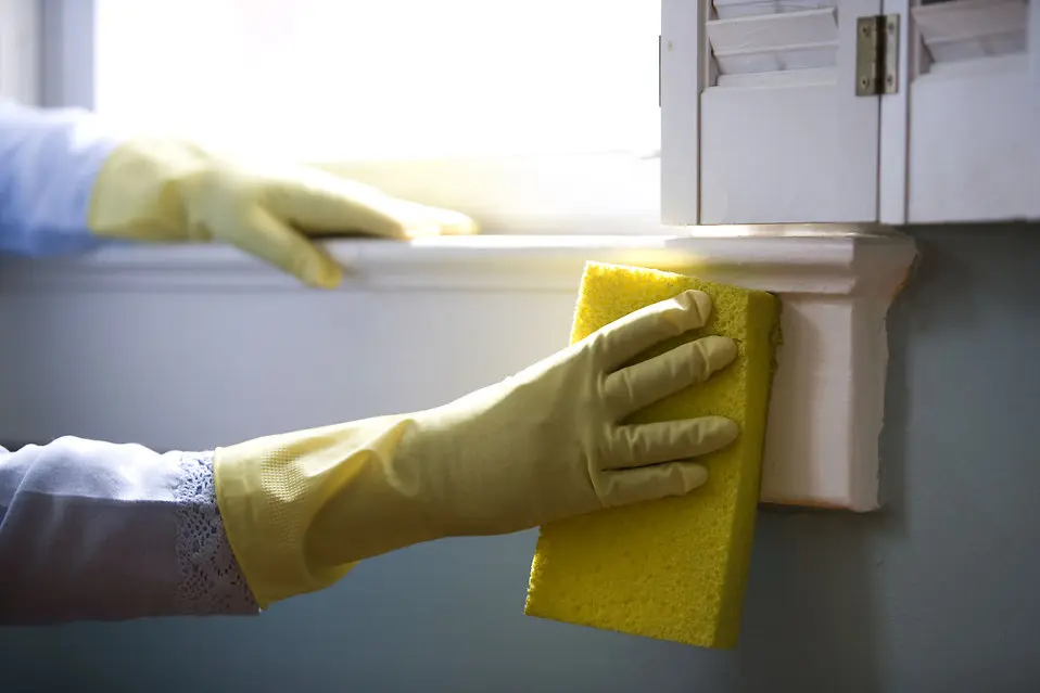 Hand wearing yellow rubber glove cleaning a window sill with a yellow sponge, illustrating home maintenance and cleanliness tips.