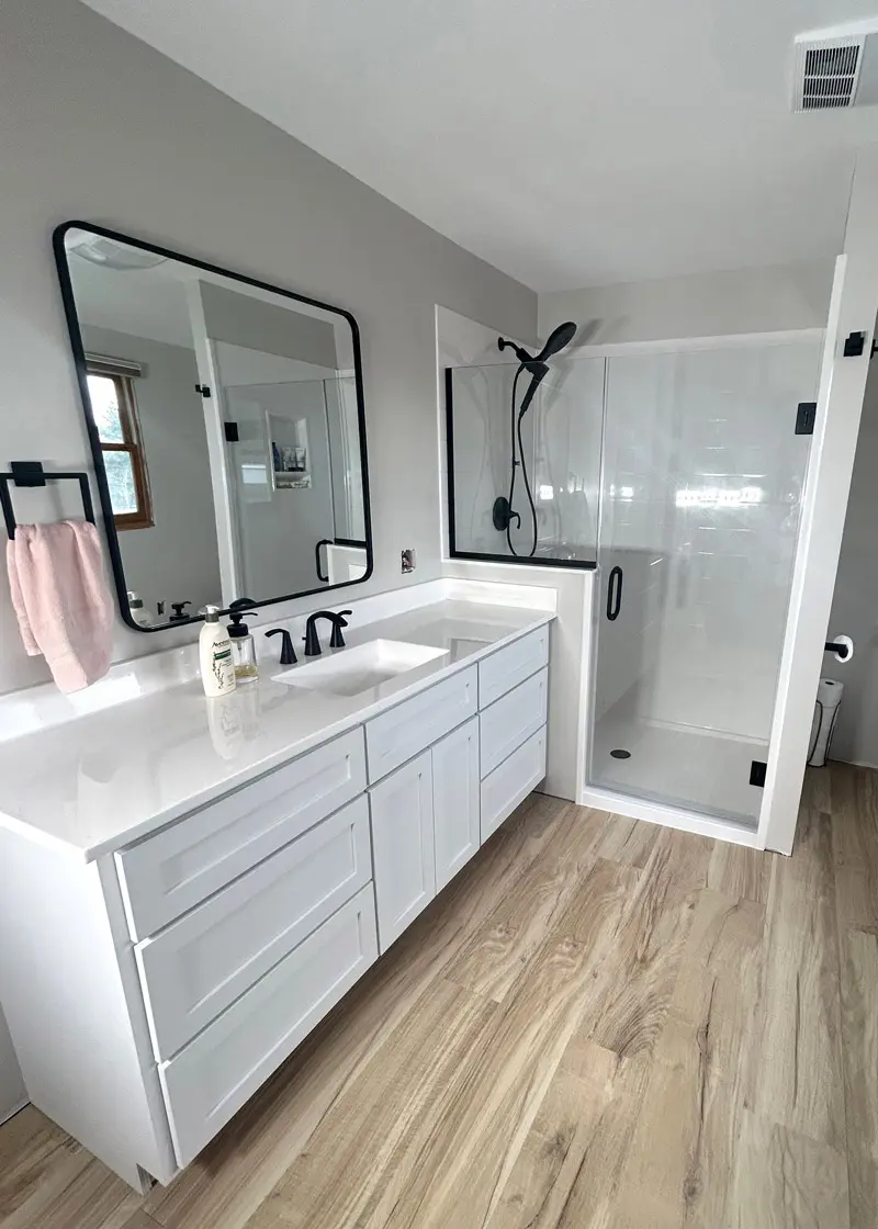 Modern bathroom featuring Onyx Collection shower system, sleek white cabinetry, black fixtures, large mirror, and wood-like flooring.