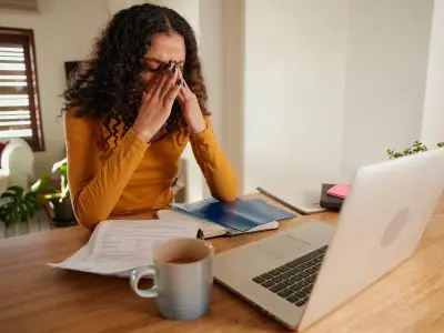 Woman with curly hair sitting at a desk, looking stressed while working on a laptop, surrounded by papers and a cup of coffee, illustrating the challenges of managing home utility costs and plumbing maintenance.