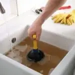 Person using a plunger to clear a clogged sink filled with brown water, illustrating plumbing issues related to root obstructions and the need for professional sewer line repair services in New York City.