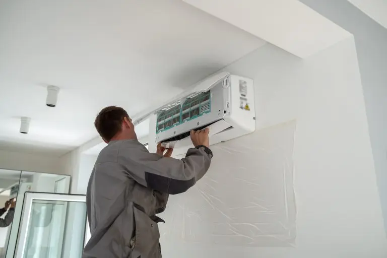 Male worker wearing uniform installing air conditioner in apartment during summer season, man technician standing indoors repairing HVAC system, checking and replacing AC filter