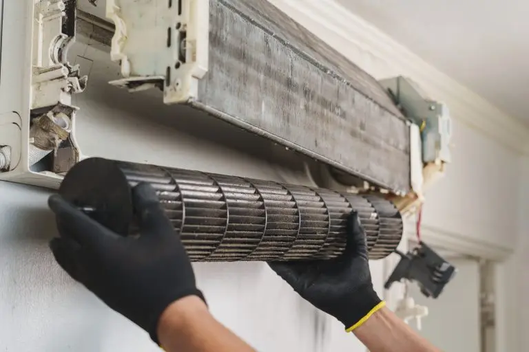 A technician is carefully removing a dirty cylindrical blower fan from an indoor wall-mounted air conditioner unit for maintenance and cleaning.