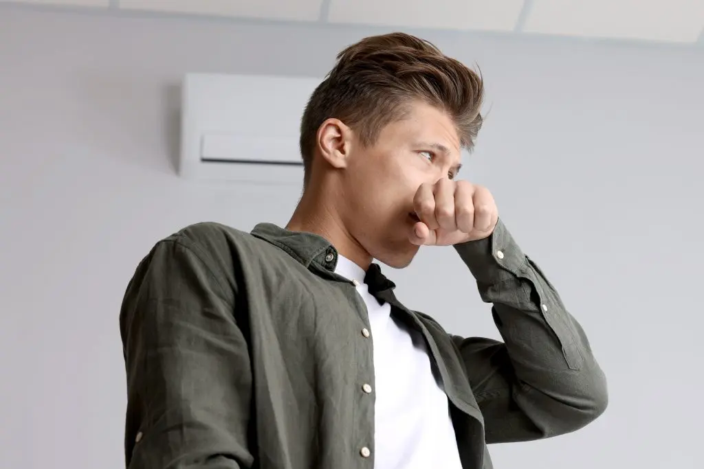Young man feeling bad smell from air conditioner indoors, low angle view