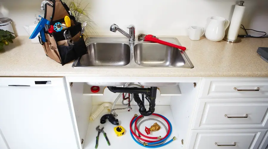 Plumbing tools and equipment under a kitchen sink, featuring a double stainless steel sink, a red plunger, and various plumbing supplies, relevant to plumbing maintenance tips for preventing holiday plumbing issues.