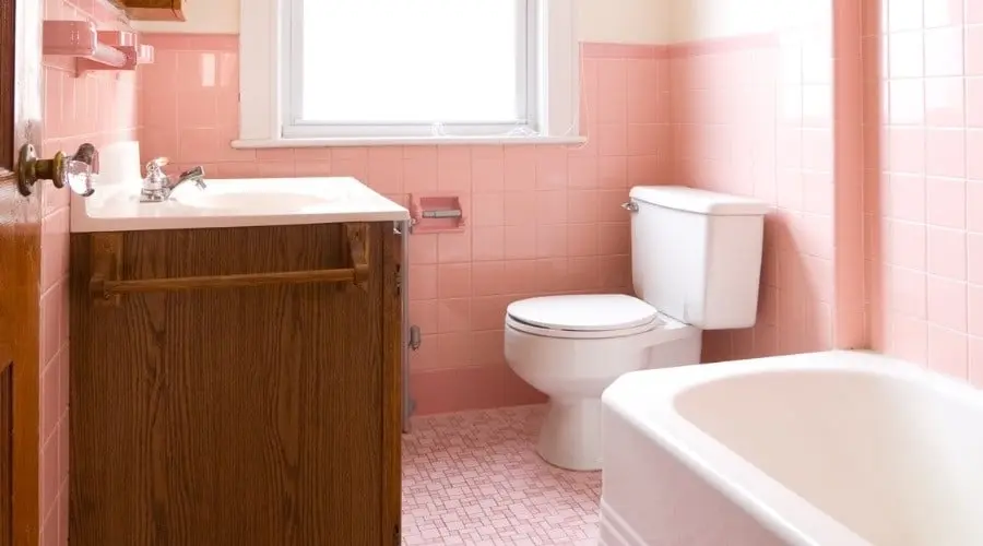 Bathroom interior featuring pink tiled walls, a wooden cabinet, a white toilet, and a bathtub, illustrating plumbing conditions in older homes.