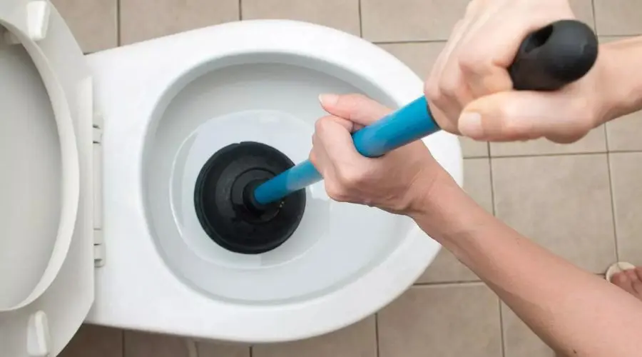 Person using a toilet plunger to address plumbing issues in a bathroom with tile flooring and a water-filled toilet bowl.