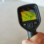 Thermal imaging camera held in hand, showcasing multicolored patterns against concrete for plumbing inspections.