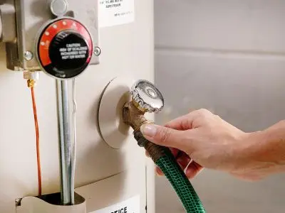 Hand connecting a hose to a water heater drain valve, illustrating the water heater maintenance process, essential for flushing and preventing sediment buildup.