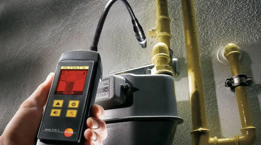 Gas leak detector held by a hand near a gas meter and pipes, featuring a digital display and flexible probe.