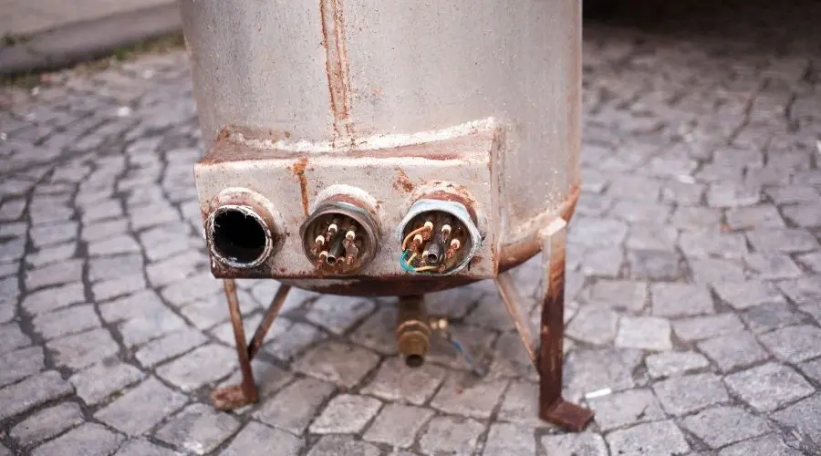 Rusted metallic heater tank with corroded surface on cobblestone floor, reflecting plumbing maintenance issues.