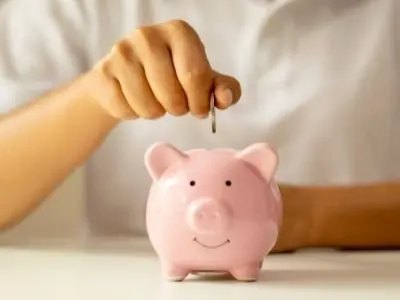 Person inserting a coin into a pink piggy bank, symbolizing saving money and financial management in the context of plumbing maintenance costs.