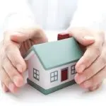 Hands protectively holding a model house with a green roof and red door, representing home ownership and plumbing care.