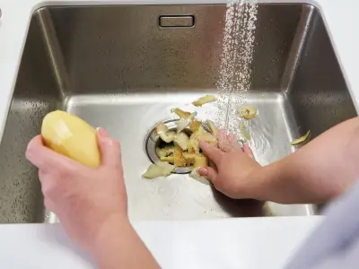 Hands rinsing vegetable peels in a kitchen sink with a garbage disposal, emphasizing proper disposal care to prevent clogs and maintain efficiency.