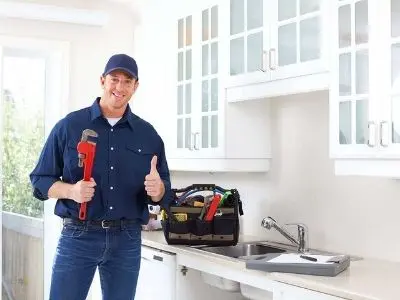 Professional plumber smiling and giving a thumbs up while holding a wrench, standing in a kitchen with tools in a bag, emphasizing plumbing services and expertise.