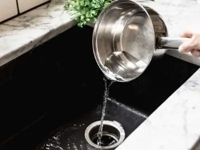 Hand pouring boiling water from a stainless steel pot into a kitchen sink drain, illustrating a method for clearing clogged drains.