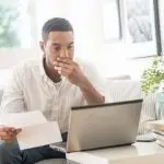 Man sitting at a desk looking concerned while holding documents and viewing a laptop, reflecting on plumbing service inquiries and issues.