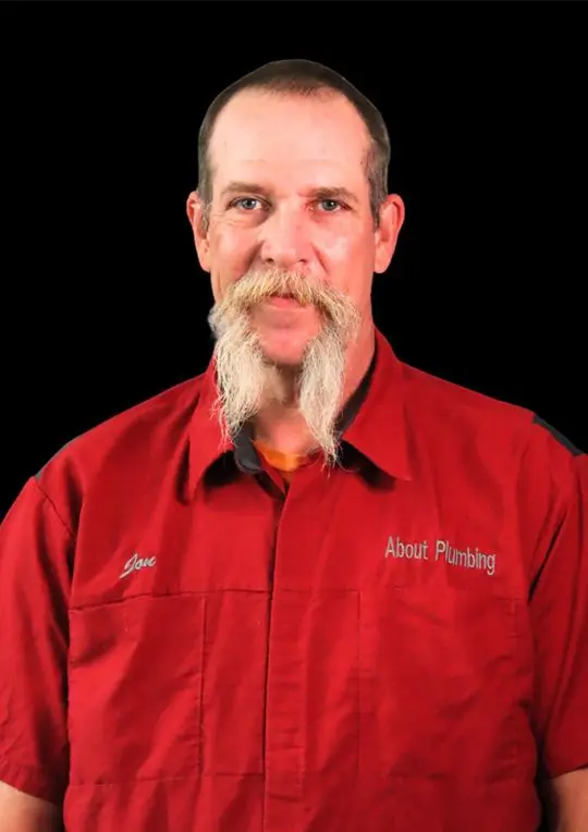 Jon Bollinger, technician at About Plumbing, wearing red uniform with company logo, smiling against a black background.