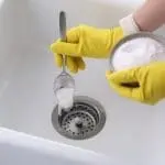 Person in yellow gloves using a spoon to add baking soda to a kitchen sink drain, illustrating effective drain cleaning methods relevant to plumbing maintenance.