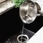 Stainless steel pot pouring water into a kitchen sink drain, highlighting plumbing maintenance and effective drain cleaning methods.