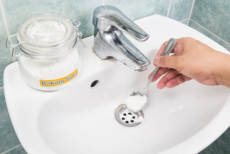 Hand pouring baking soda from a spoon into a bathroom sink drain, with a jar labeled "Baking Soda" nearby, illustrating a DIY method for unclogging sinks.
