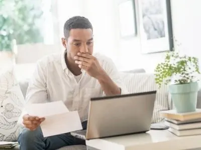 Man looking concerned while holding a water bill and using a laptop, illustrating issues related to underground plumbing leaks and rising utility costs.