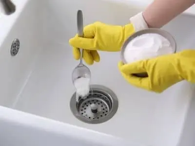 Person wearing yellow gloves pouring baking soda into a kitchen sink drain, demonstrating a method for clearing clogs using baking soda and vinegar.