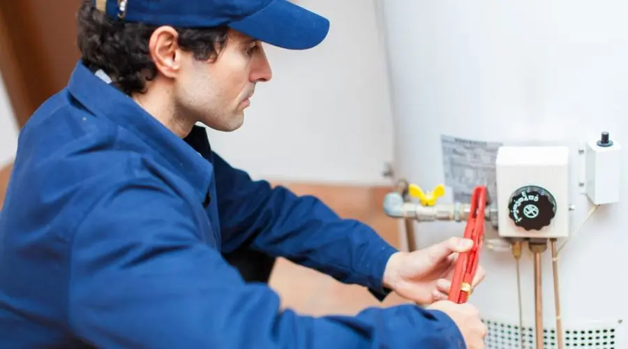 Male technician in blue uniform repairs water heater with pipe wrench and control panel. Hancock & Son Plumbing services.