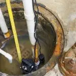 Photo of a sump pump setup with water accumulation, PVC pipes, and measurement tape by Hancock & Son Plumbing.
