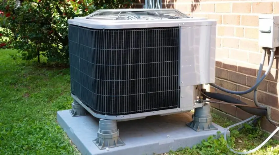 Air conditioning unit outdoors on concrete slab, featuring metallic casing, hoses, and nearby grass and brick wall.
