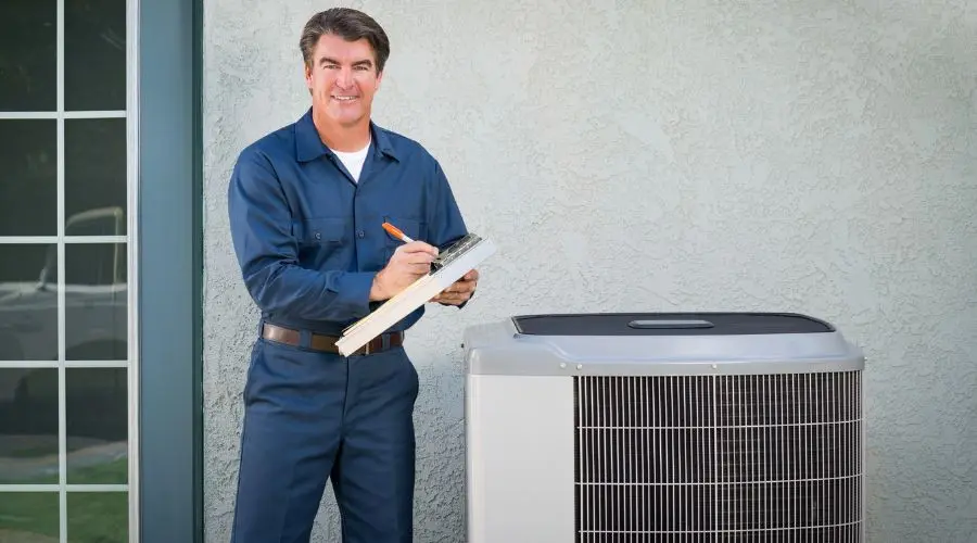 Man in technician attire with clipboard next to air conditioning unit, highlighting HVAC service by Hancock & Son Plumbing.