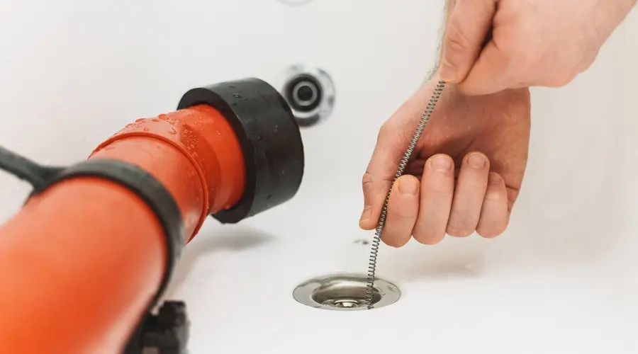 Person using a plumbing snake to clear a drain, featuring a red pipe and sink, relevant to drain cleaning services and sewer line maintenance.