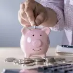 Pink piggy bank with a smiling face, a hand inserting a coin, and scattered coins, representing savings for HVAC efficiency.