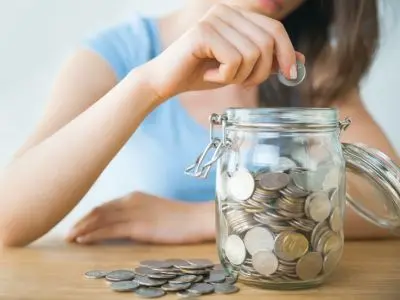 Person adding a coin to a glass jar filled with coins, symbolizing savings and financial efficiency related to air conditioning maintenance costs.