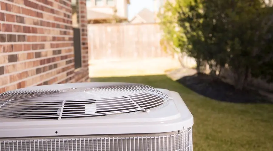 Air conditioning unit on a brick house exterior with lawn, featuring a metal grill and fan, in a residential outdoor setting.