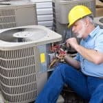 Older male technician working on air conditioning units for AirLux Heating & Cooling, wearing a hard hat and blue shirt.
