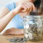 Person depositing a coin into a glass jar filled with coins, symbolizing financial responsibility and savings.